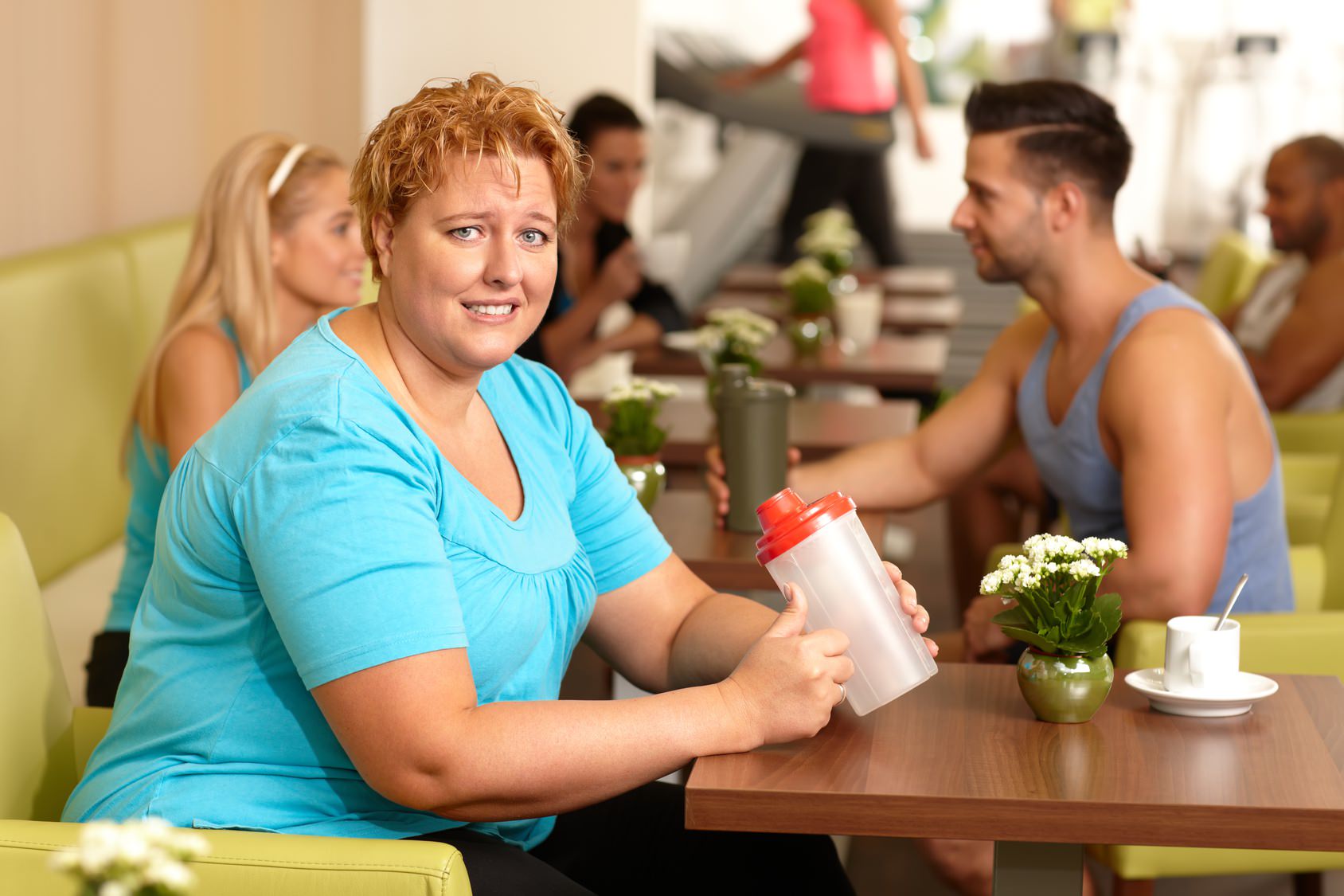 Übergewichtige Frau sitzt im Fitnessstudio und beäugt einen Shake sehr kritisch